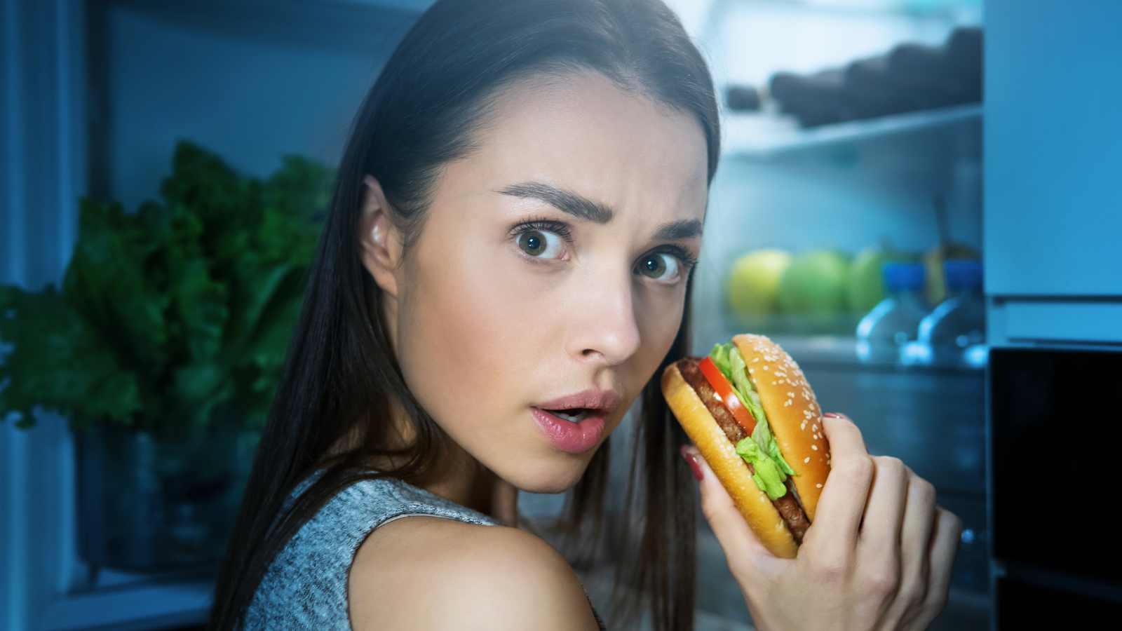 young woman eating burger from fridge